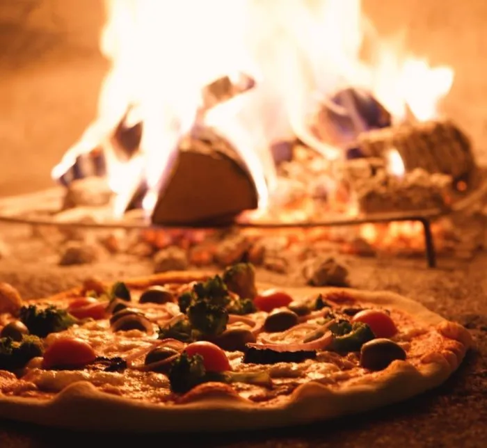 Pizza mit Gemüse im Holz-Steinofen, dahinter das offene Feuer